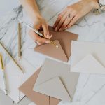 Flat lay of hands writing on envelopes with pencils on marble surface.